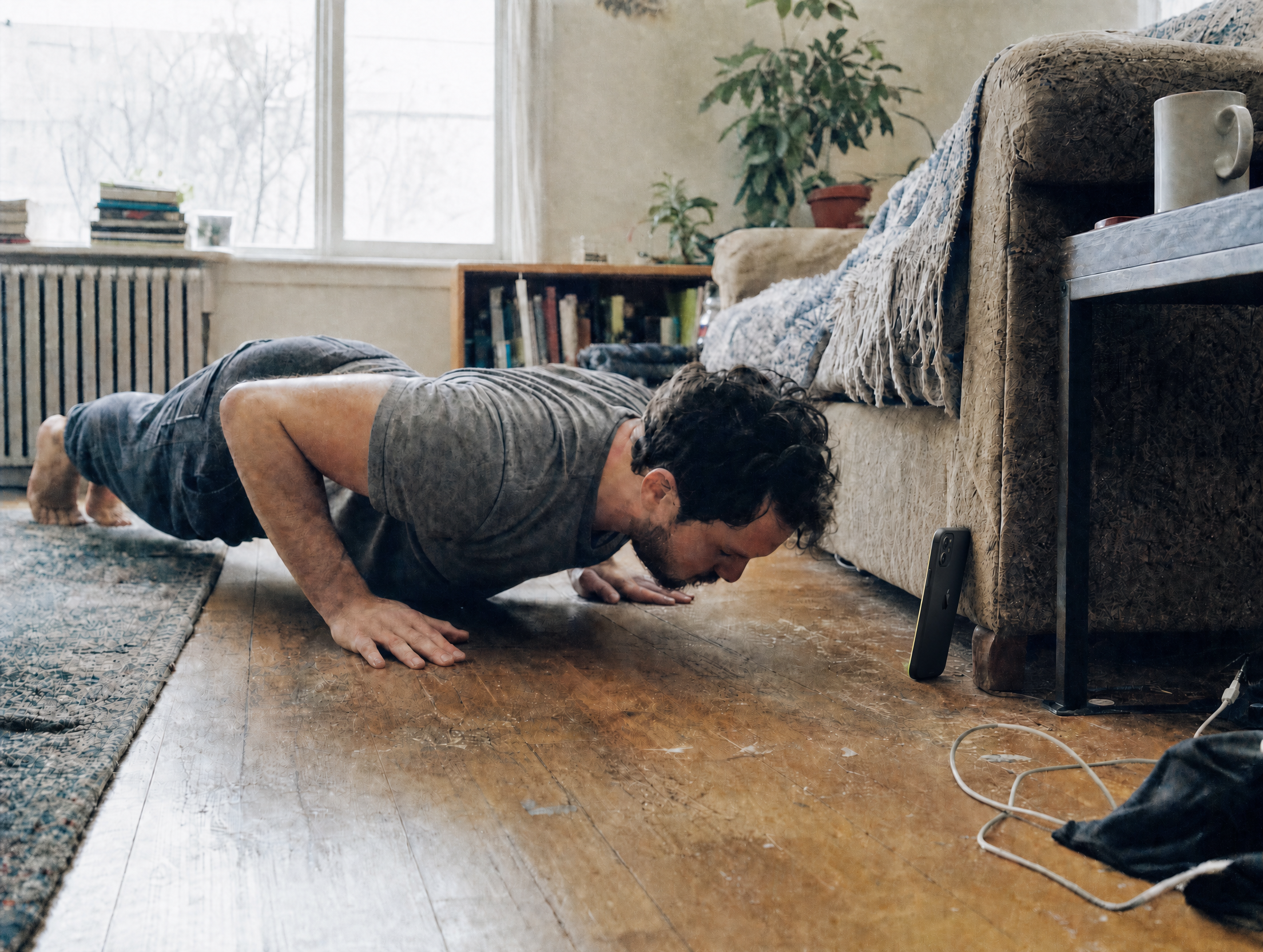 A man mid-pushup on a living room floor, an iPhone propped against the front edge of the couch with its screen turned toward his face.